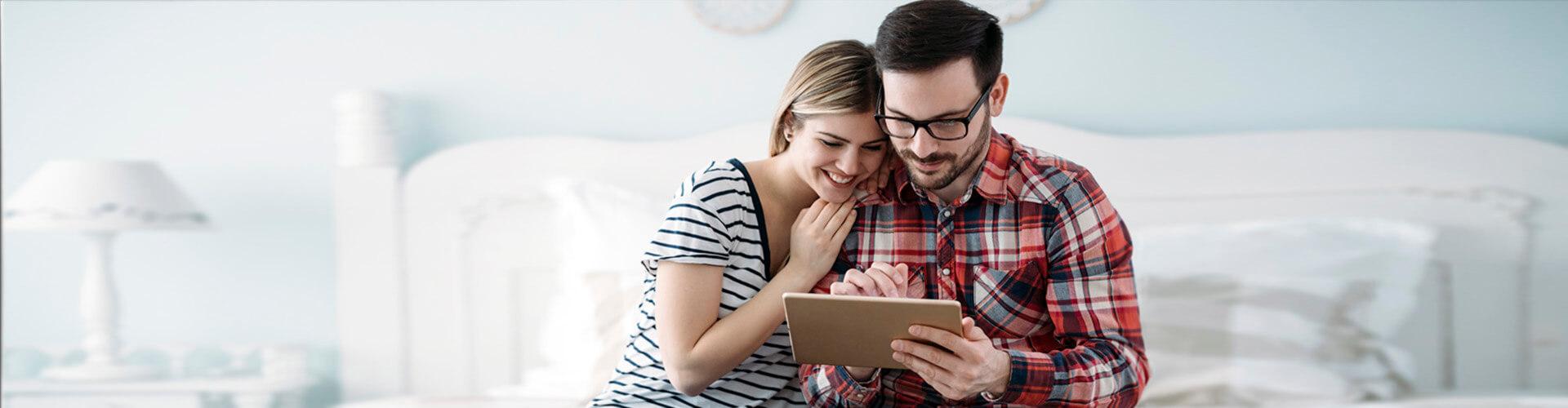 Programa te lo mereces nomina y pension - Pareja sentada en su cama en la habitacion con la tablet en la mano sonriendo