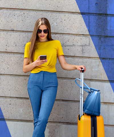 Momento Viajar al extranjero - mujer-joven-con-camiseta-amarilla-y-maleta-usando-su-movil-apoyada-contra-una-pared-azul-y-gris - Rural Kutxa