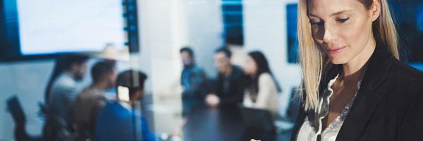Seguros para Empresas - Mujer joven y rubia de negocios con chaqueta de traje negra y camisa blanca utilizando una tablet en una reunion en la oficina Seguros para Empresas - Mujer joven y rubia de negocios con chaqueta de traje negra y camisa blanca utilizando una tablet en una reunion en la oficina